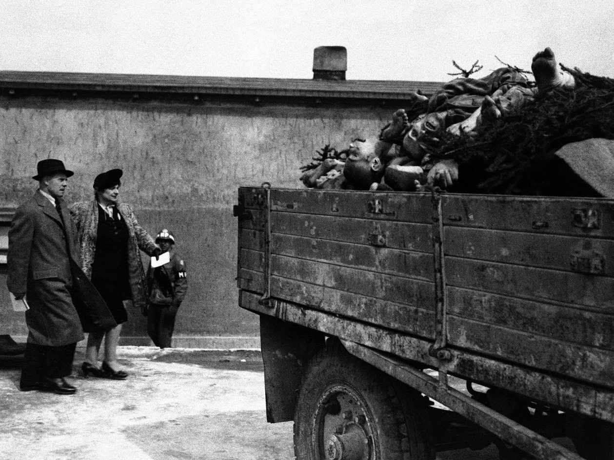 Eine historische Schwarz-Weiß-Fotografie aus dem Konzentrationslager Buchenwald 1945. Rechts im Bild steht ein Lastwagen mit offener Ladefläche, auf dem zahlreiche Leichen ermordeter KZ-Häftlinge übereinandergestapelt liegen. Links im Vordergrund gehen zwei Mitglieder einer britischen Parlamentsdelegation – ein Mann im Mantel und Hut sowie eine Frau im Kostüm und Hut – an dem Fahrzeug vorbei. Im Hintergrund ist ein alliierter Soldat in Uniform zu sehen, der die Szene beobachtet. Das Bild dokumentiert die unmittelbare Konfrontation von Politikern mit den Verbrechen des NS-Regimes.