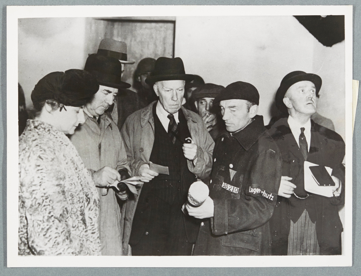 Eine historische Schwarz-Weiß-Fotografie zeigt Mitglieder einer britischen Parlamentsdelegation beim Besuch des Konzentrationslagers Buchenwald im April 1945. Links steht eine Frau in Mantel und Hut, neben ihr mehrere Männer in Anzügen und Mänteln mit Hüten, die aufmerksam zuhören und Notizen machen. Im Vordergrund hält ein KZ-Häftling in einer Jacke mit der Aufschrift „Lagerschutz“ und der Häftlingsnummer „88210“ einen Gegenstand in den Händen, während er spricht. Die Delegierten blicken ernst und konzentriert auf die Szene.