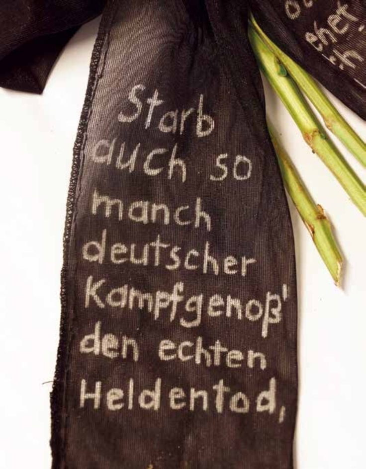 Aufschrift Blumengebinde: Starb auch so manch deutscher Kampfgenoß' den echten Heldentod, [...].