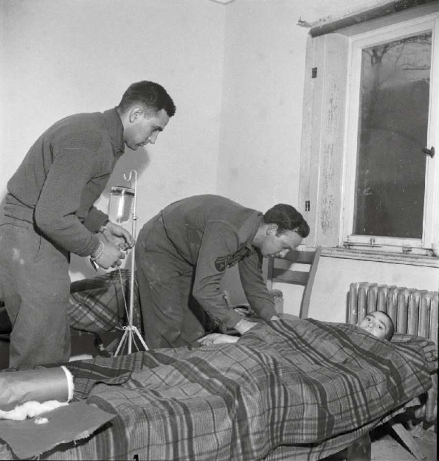 Schwarz-Weiß-Foto: zwei amerikanische Sanitäter versorgen einen Überlebenden, der zugedeckt in einem Bett liegt