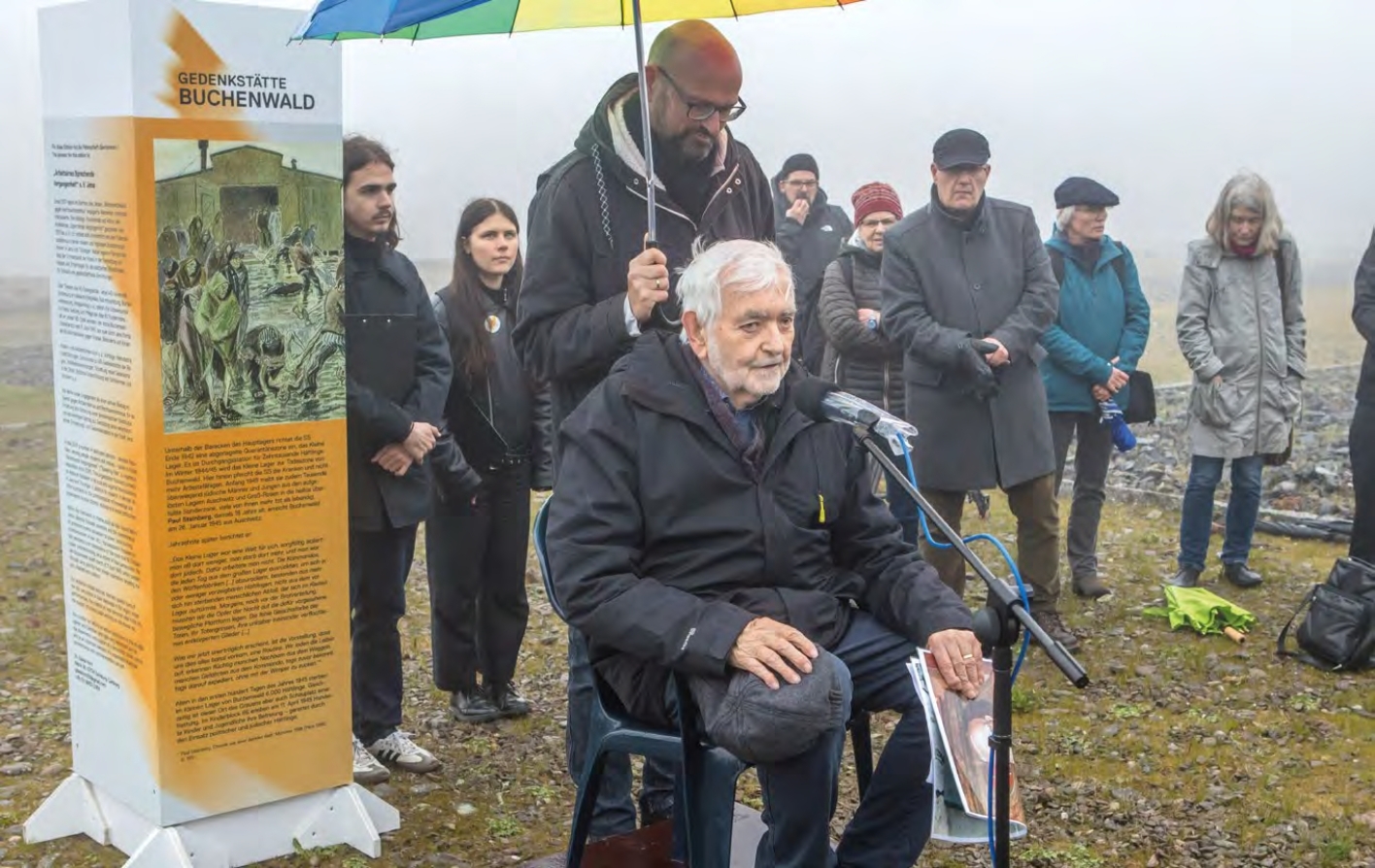 Wolfgang Rug sitzt vor einem Mikrofon und spricht zu der Menge auf dem Lagergelände Buchenwalds