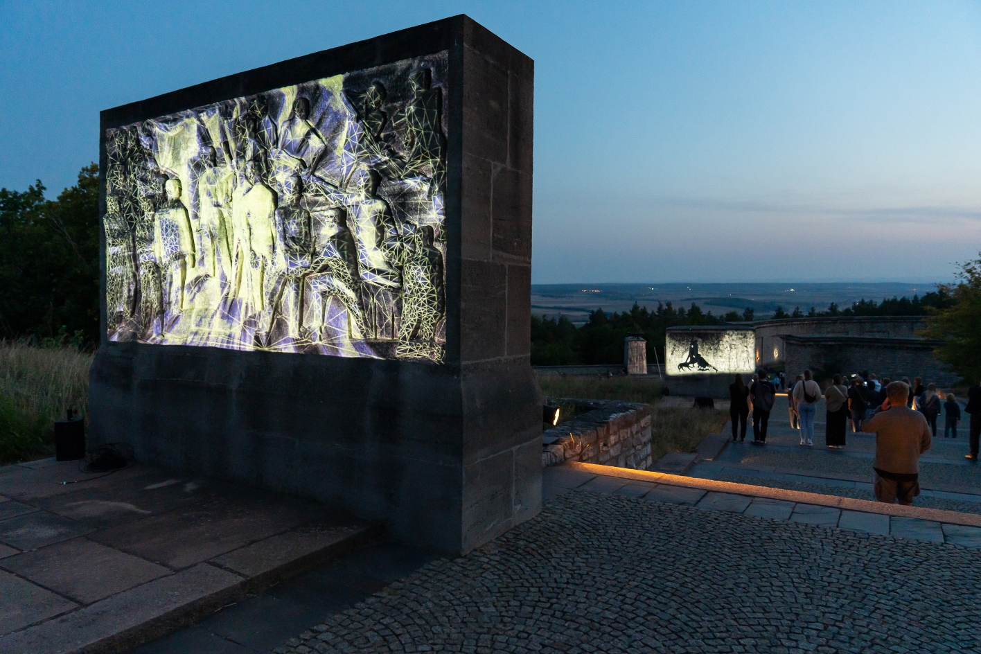 Abendliche Szene an einem Denkmal mit mehreren steinernen Reliefs, auf die digitale Lichtprojektionen mit grafischen Mustern und Figuren geworfen werden. Besucher:innen gehen die Treppen entlang und betrachten die Kunstinstallation in der Dämmerung.