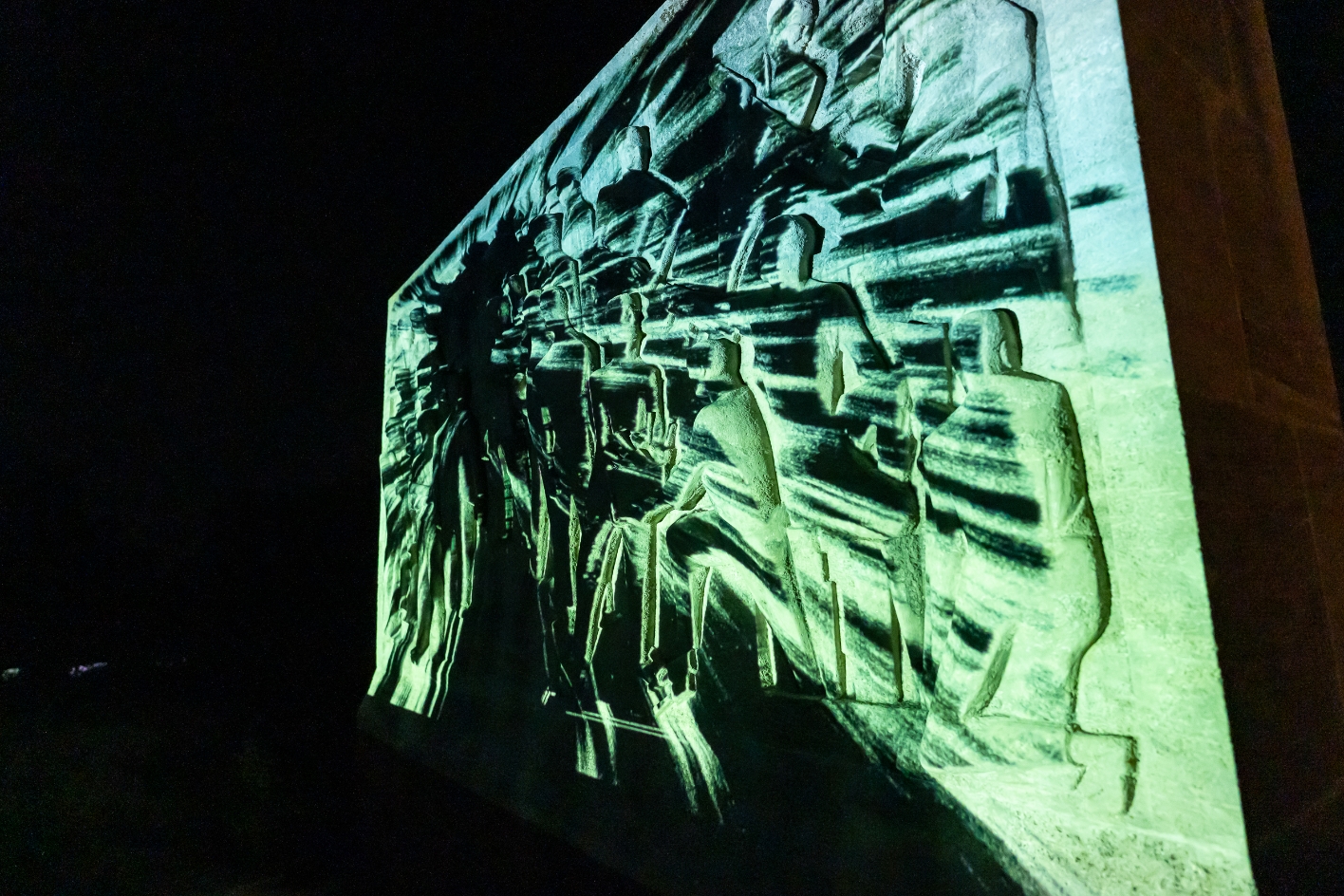Reliefwand bei Nacht, auf die mit grünlich-weißen Lichtstrahlen abstrakte Muster projiziert werden. Die Darstellung überlagert die menschlichen Figuren des Denkmals mit verzerrten Linien und schafft eine dynamische, fast bewegte Wirkung im dunklen Umfeld.
