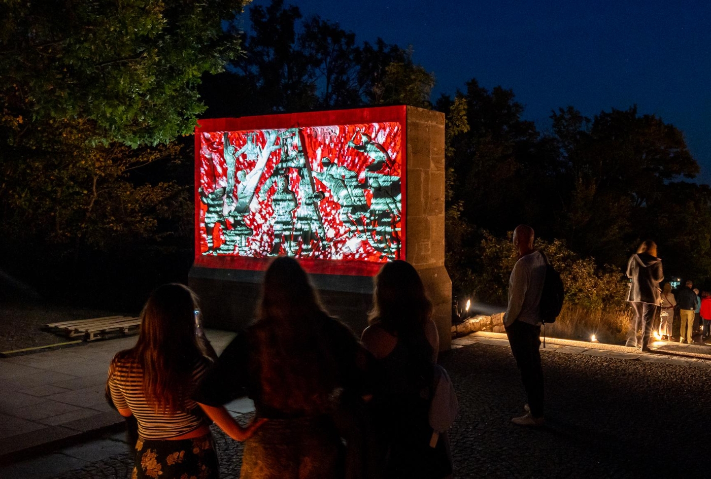 Besucher:innen betrachten bei Nacht eine bunte Videoprojektion auf einem Denkmal am Mahnmal Buchenwald während des Genius Loci Festivals 2024; im Hintergrund leuchtet das Lichtermeer der Stadt Weimar
