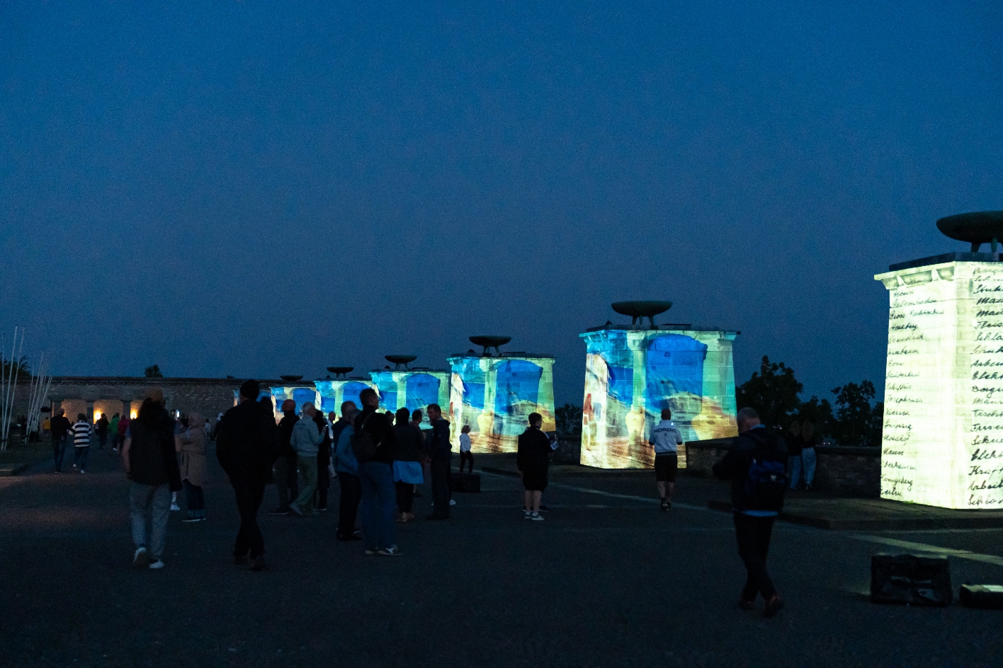 Abendliche Projektion beim Genius Loci Festival 2024 auf den Nationensteinen des Buchenwald-Mahnmals: Menschen betrachten die mit farbigen Animationen und Schriftzügen beleuchteten Gedenksäulen