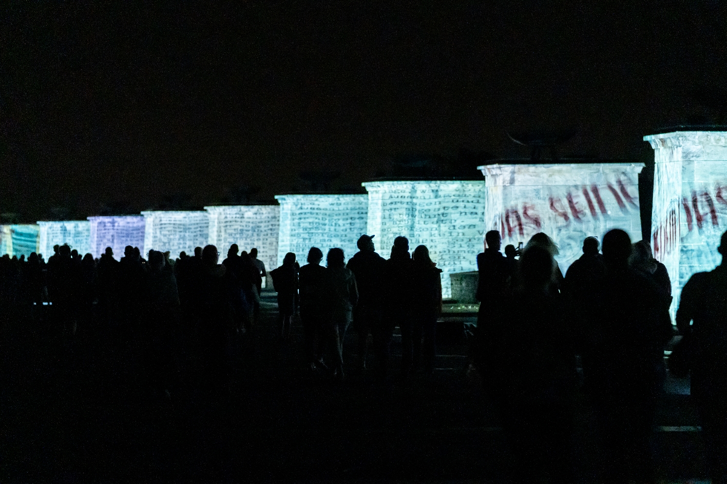 Besucher:innen betrachten bei Nacht die Nationensteine des Buchenwald-Mahnmals, auf die beim Genius Loci Festival 2024 bewegte Text- und Bildprojektionen geworfen werden – darunter große rote Schriftzüge wie ‚Du das Sein‘