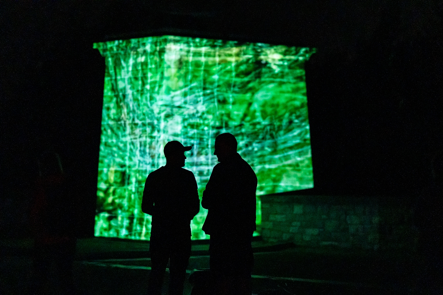 Zwei Personen stehen bei Nacht silhouettenhaft vor einem grün illuminierten Nationenstein des Buchenwald-Mahnmals, auf den beim Genius Loci Festival 2024 eine abstrakte Netzstruktur projiziert wird.