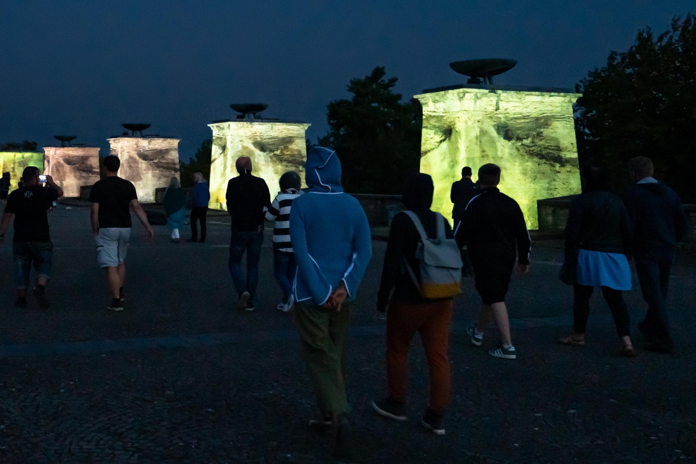 Besucher:innen gehen in der Dämmerung über das Gelände des Buchenwald-Mahnmals, während die Nationensteine durch farbige Projektionen im Rahmen des Genius Loci Festivals 2024 illuminiert werden.