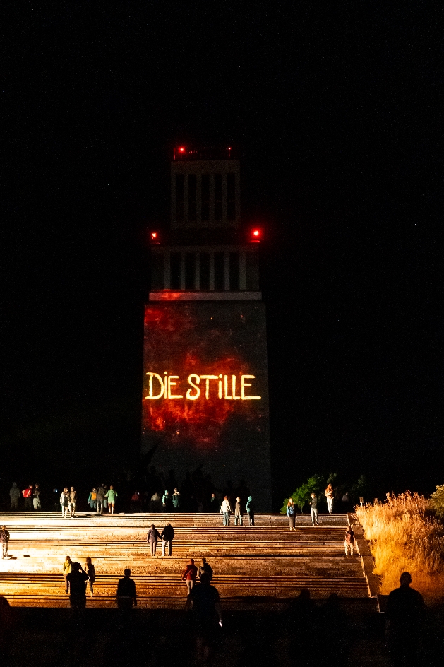 Nächtliche Szene am Glockenturm der Gedenkstätte Buchenwald: Menschen steigen die beleuchtete Treppe hinauf, während auf dem Turm in leuchtenden Buchstaben die Worte ‚DIE STILLE‘ projiziert werden.