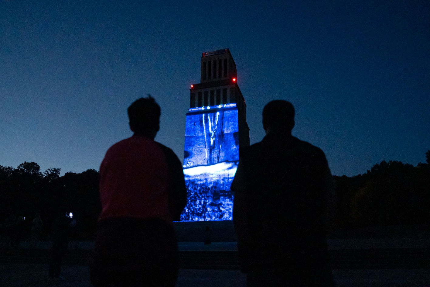 Zwei Personen im Vordergrund blicken bei Dunkelheit auf den beleuchteten Glockenturm der Gedenkstätte Buchenwald, auf dem eine blaue Lichtprojektion mit abstrakten Formen zu sehen ist.