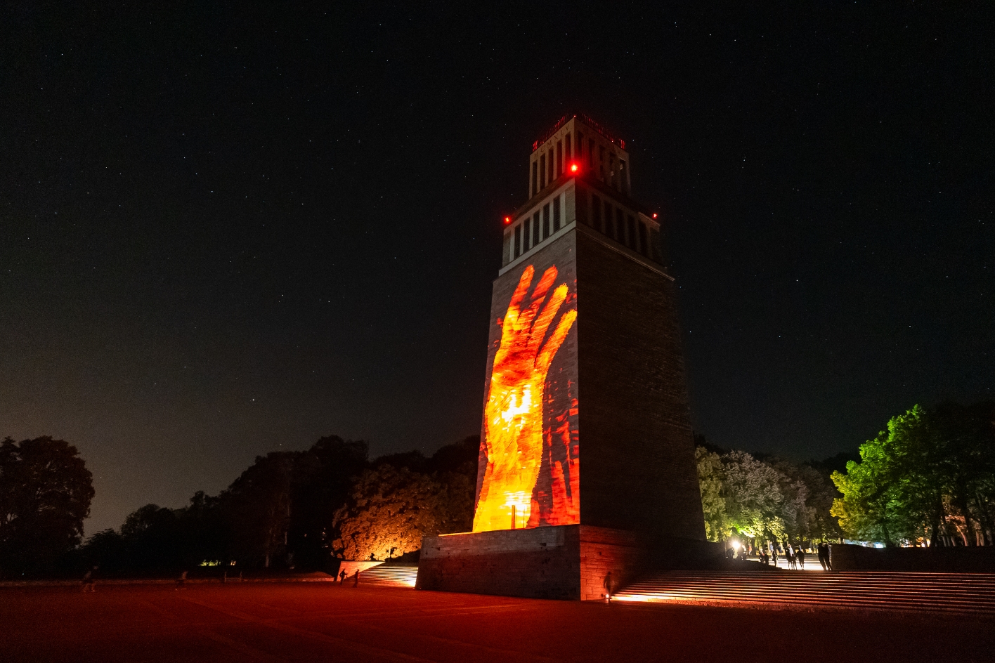 Nachtaufnahme des Glockenturms der Gedenkstätte Buchenwald mit einer rot-orangenen Lichtprojektion einer erhobenen Hand auf der Turmfassade, unter einem klaren Sternenhimmel.