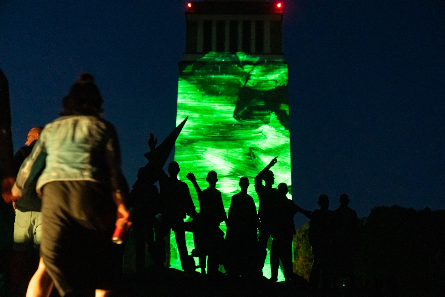 Menschen vor dem Glockenturm der Gedenkstätte Buchenwald bei Nacht, im Vordergrund die silhouettenhafte Figurengruppe des Denkmals; auf den Turm wird eine abstrakte grün leuchtende Lichtprojektion geworfen.
