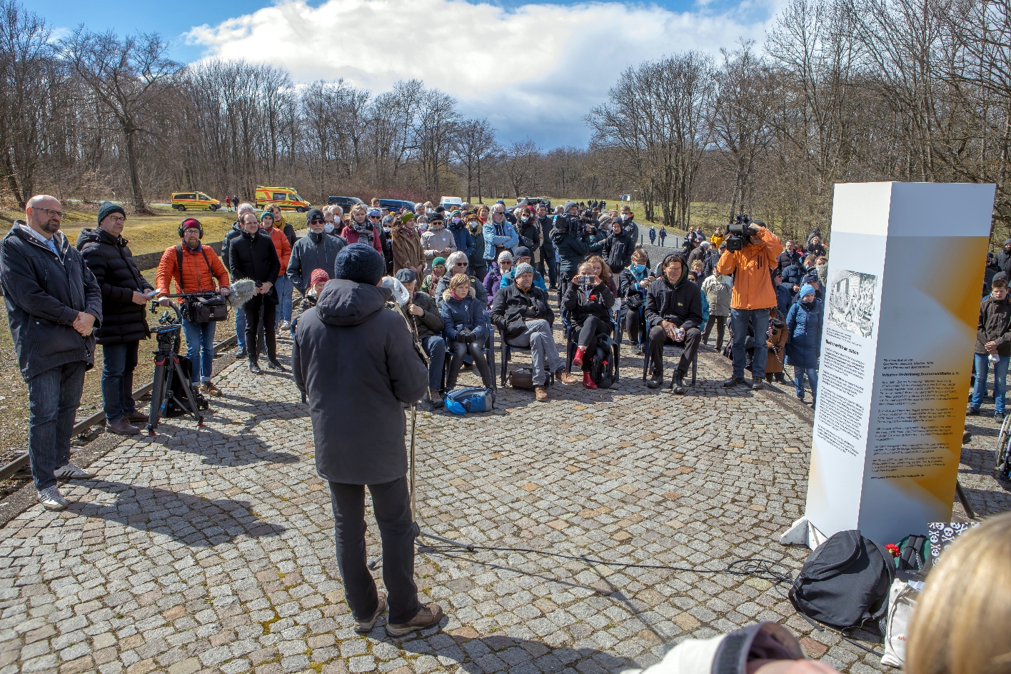 In the foreground, a speaker with a green jacket and blue cap can be seen from behind, speaking into a microphone. To the right of him stands a gray-white-orange information pillar, which offers information about prisoner transports. In the background, a crowd of people can be seen, which has gathered in front of the speaker with some distance to him.