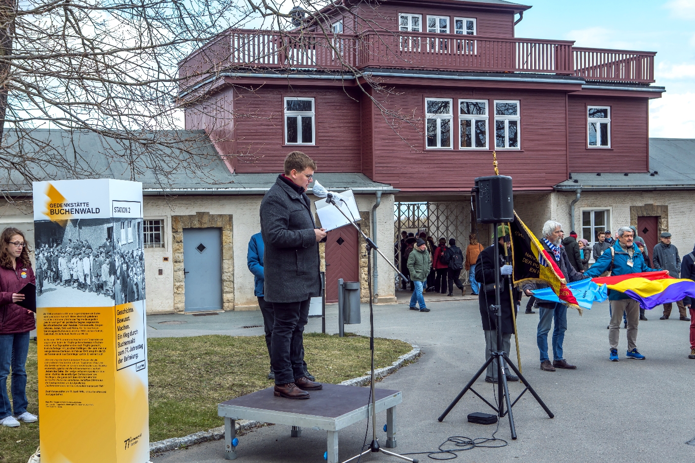 Im Zentrum des Bildes steht ein Redner mit schwarzer Jacke auf einem kleinen Bühnenelement und spricht in ein Mikrofon. Links neben ihm steht eine grau-weiß-orangene Info-Stele. Im Hintergrund ist das Torgebäude des ehemaligen Konzentrationslagers zu sehen.