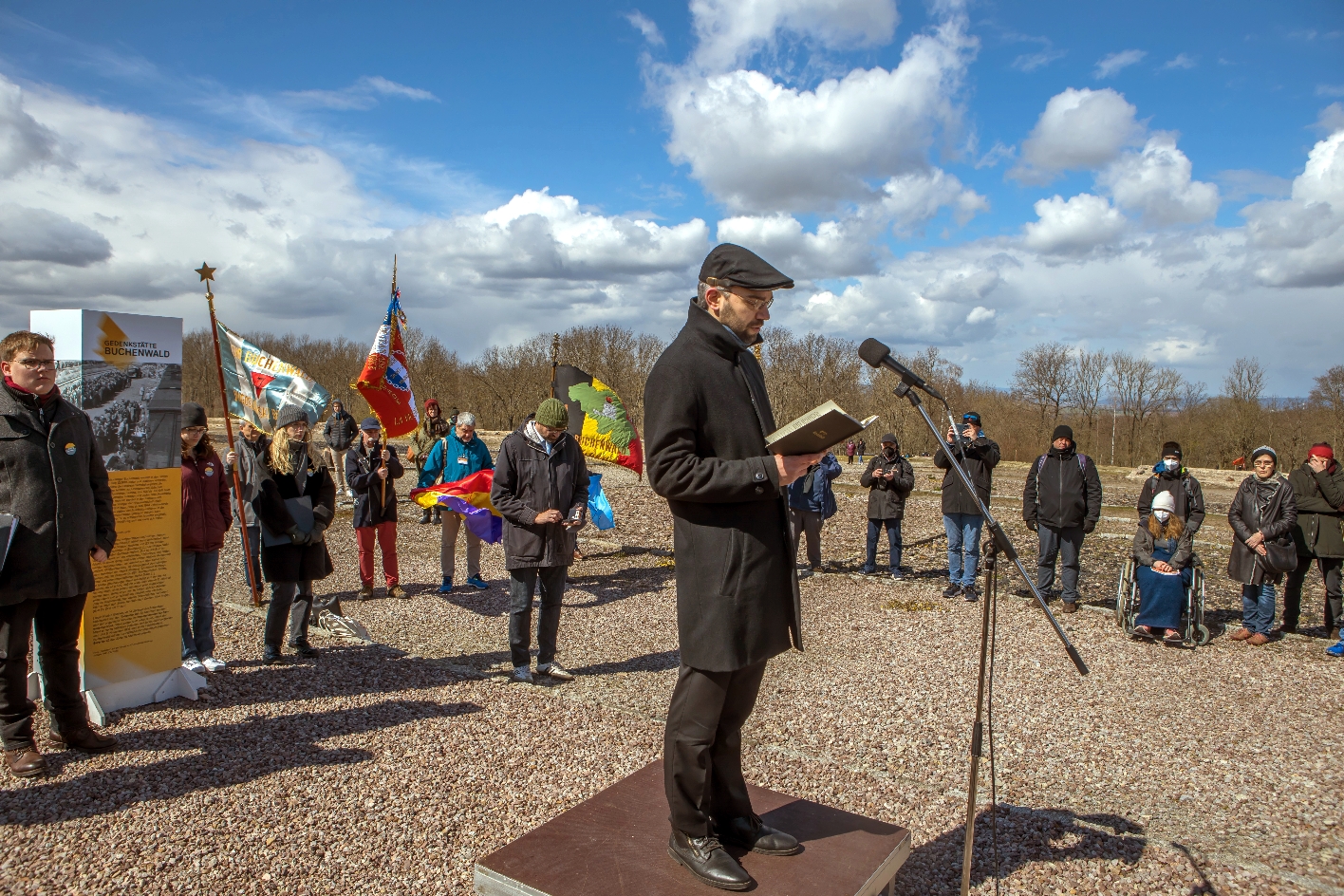 In the center of the picture the national rabbi Alexander Nachama stands on a small stage element and speaks into a microphone. In the background are listeners, some with flags.