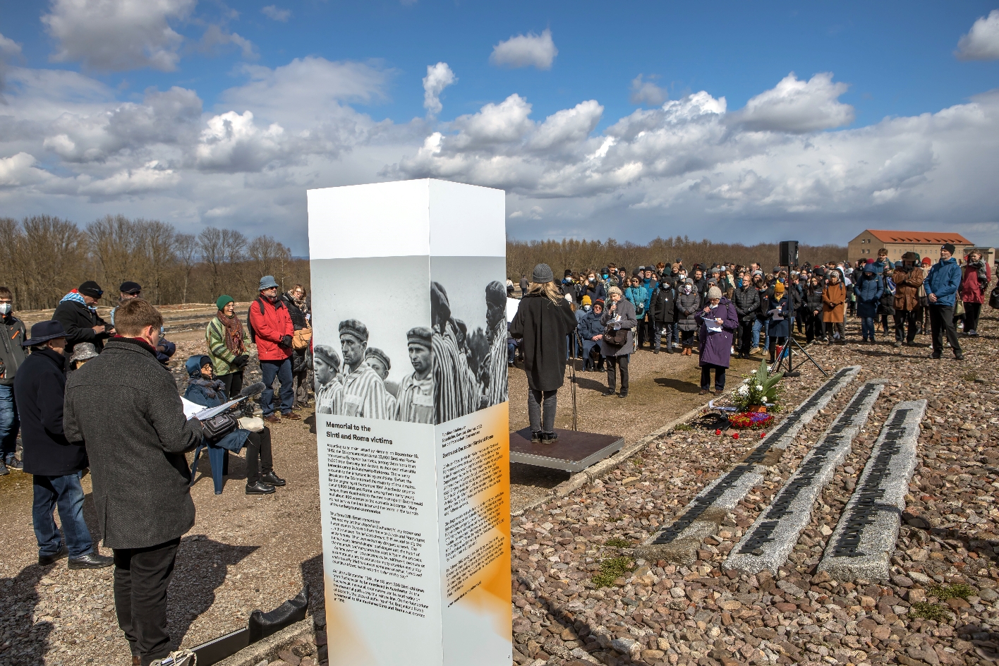 Im Vordergrund ist eine weiß-grau-orange Infostele zu sehen, auf der Erklärungen in verschiedenen Sprachen angeboten werden. Des Weiteren sind von allen Seiten Ausschnitte einer Fotografie von Burgenländer Roma in Häftlingskleidung zu sehen. Links neben der Stele steht ein Redner an einem Mikrofon. Im Hintergrund ist eine Menschenmenge zu sehen die in Richtung des Redners blickt.