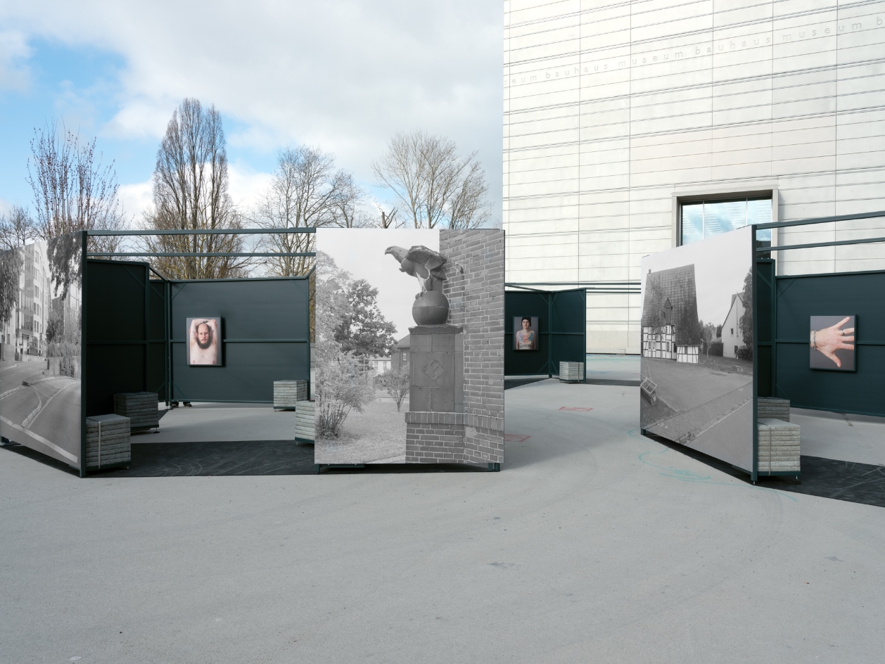 In a public square there are metal boxes with photos hanging on their walls. On the outsides are large photos of buildings in Weimar in the 40s, inside photos of people showing their tattoos. 