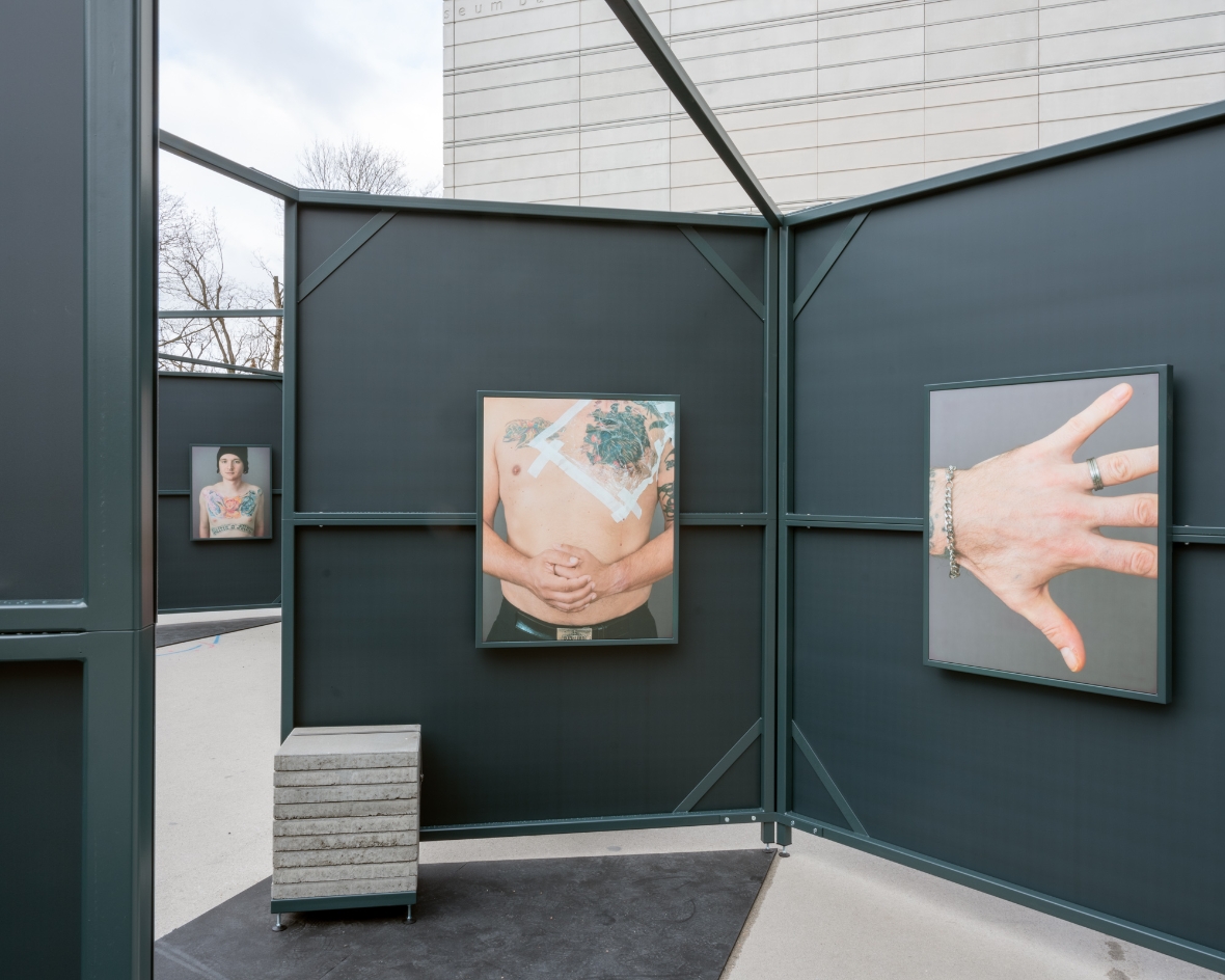 The picture shows one of the exhibition cells from the inside. Inside hang two pictures. The picture on the right shows a hand. The picture on the left shows the naked belly of a man with tattoos and patches on his chest. 