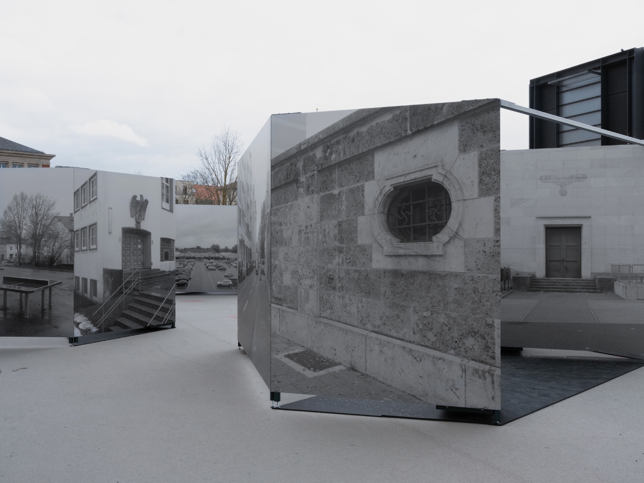 Metal boxes are placed in a public square. On the outer walls are large photos of facades and buildings from the Weimar of the 40s. The imperial eagle can be seen on some of them.
