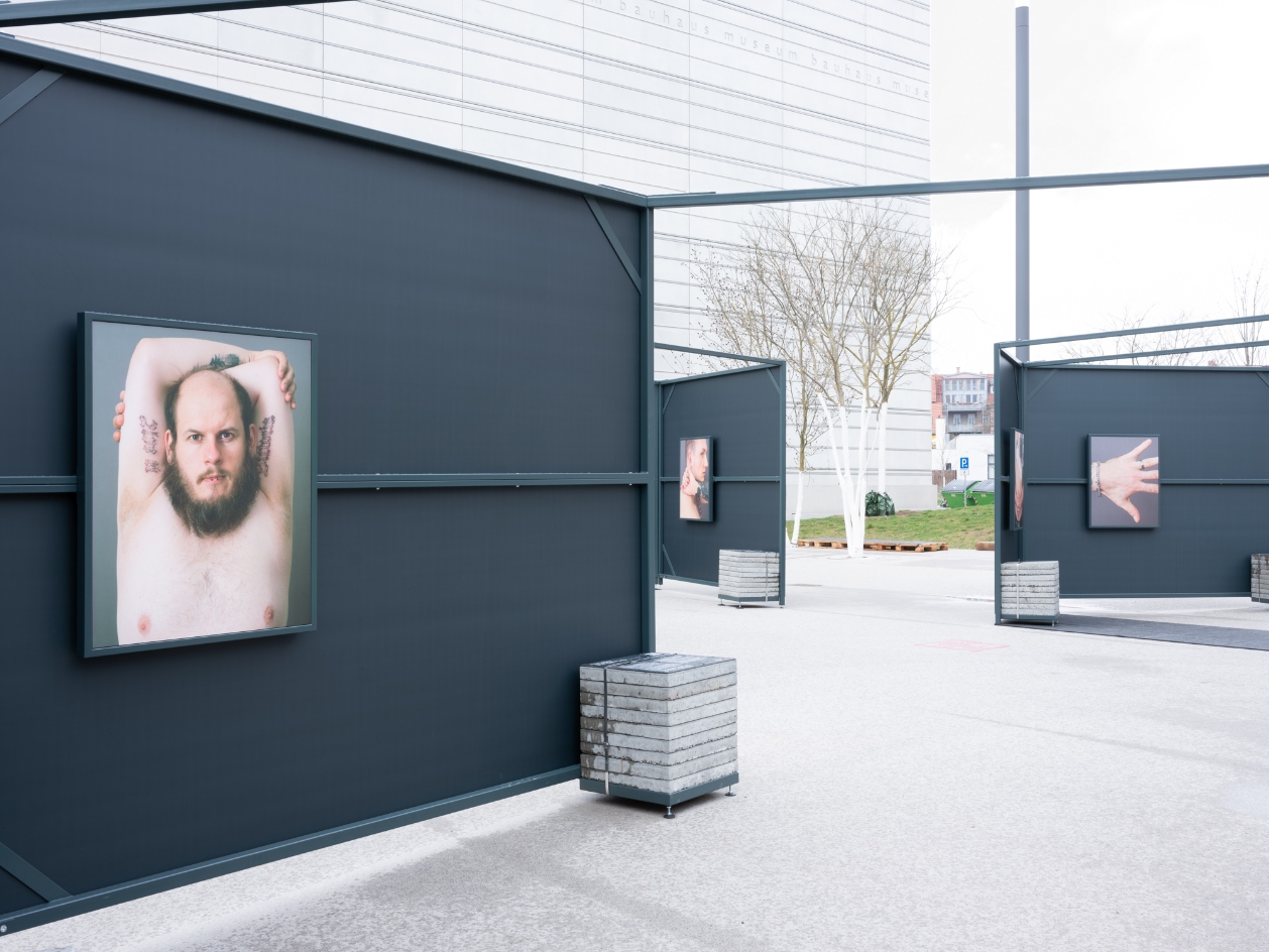 In a public square, metal boxes are set up with photos hanging on their walls. In the foreground is the image of a topless man with a beard and bald head. In the back two exhibition cells are pictures of a hand and a man in profile view.