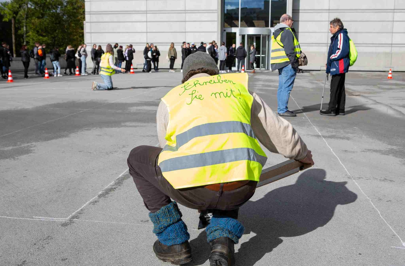 A man kneels on a stone square and wears a yellow high-visibility vest with the inscription "Write with" on his back. In his hands he holds a sticky board, which he puts straight in front of him. In the background, other people with high-visibility vests are standing and kneeling in the square.