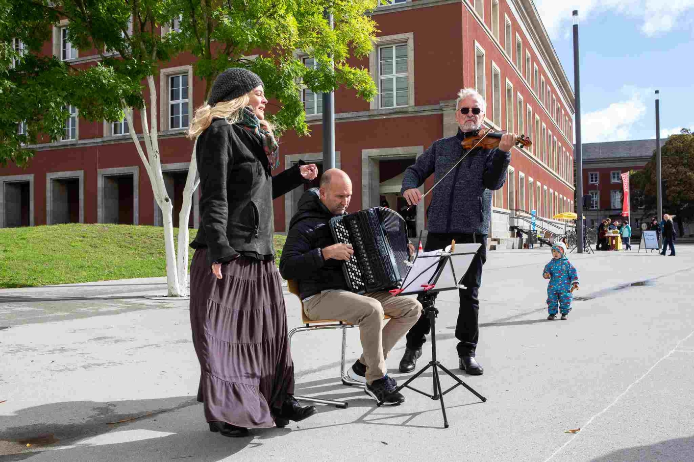 A violinist, an accordion player and a singer of the String Company Erfurt accompany the writing action musically. In the background, a toddler in a blue anorak can be seen standing free in the square and looking eagerly at the musicians.