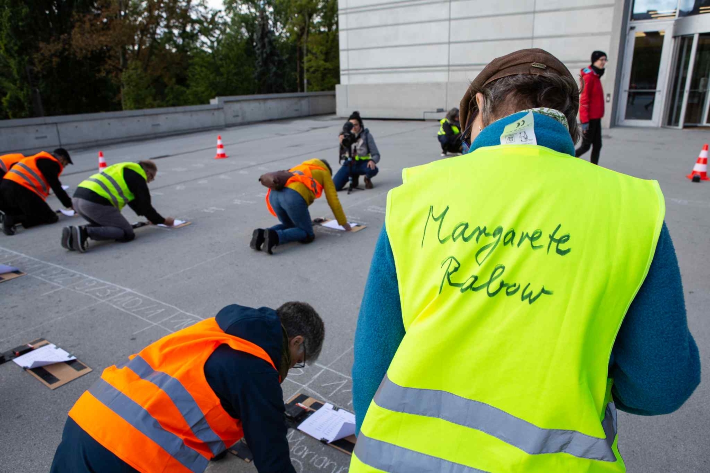 The initiator of the writing campaign, Margarete Rabow, instructs volunteer writers. She can be recognized by a warning vest with her name on the back. In the background, many participants kneel in orange vests and write their names on the ground with chalk.