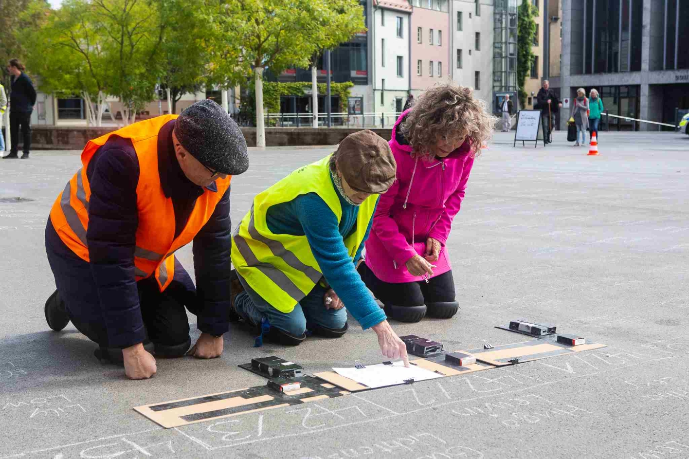 The Lantag president Birgit Pommer kneels on the left and wears a pink anorak. Margarete Rabow kneels to her left and wears a yellow high-visibility vest. In front of them on the floor stencils and chalk writing.