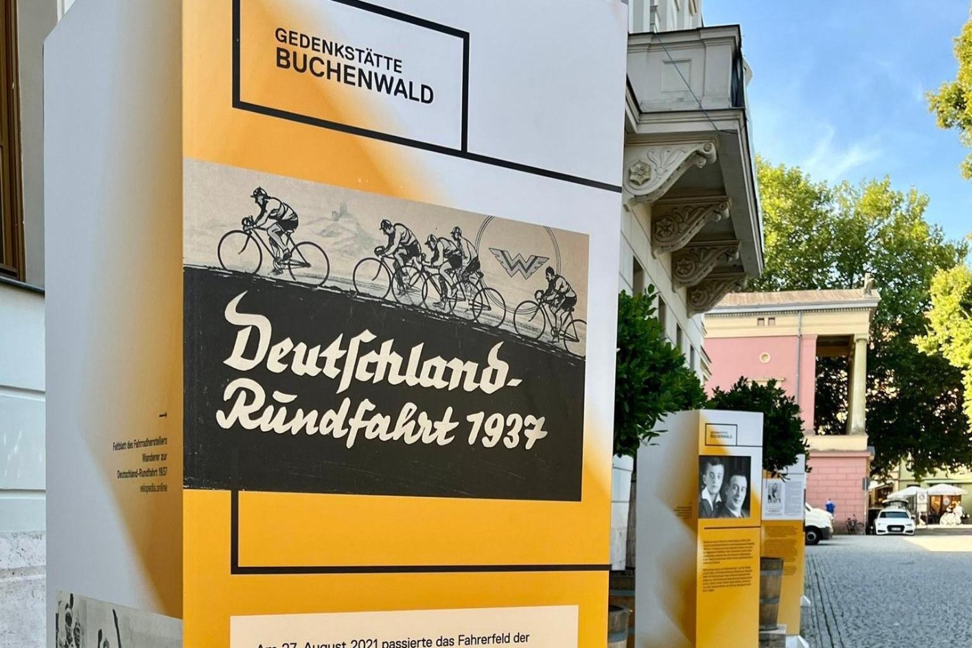 Eine weiß-orangene Infostele der Gedenkstätte Buchenwald, auf der ein Schild der "Deutschlandrundfahrt 1937" abgebildet ist. Darunter wird erklärender Text angeboten.