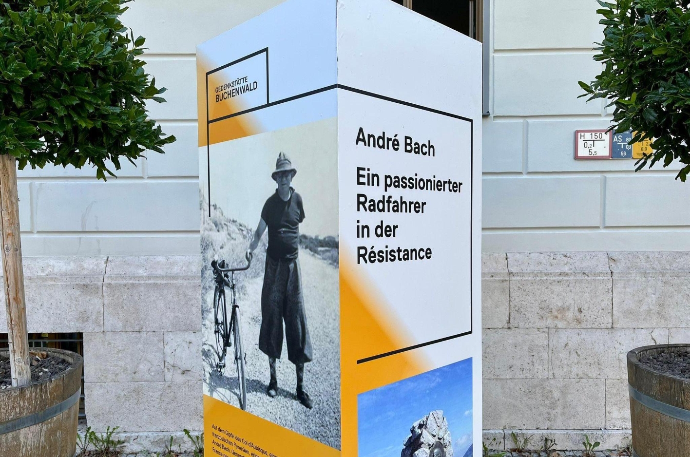 2 sides of a cuboid white-orange info-stele on which is a picture of a man next to his bicycle. On another side the information is offered that it is the member of the Resistance André Bach.