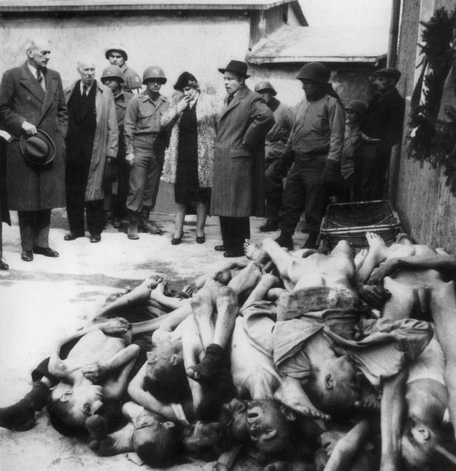 Eine historische Schwarz-Weiß-Fotografie aus dem Konzentrationslager Buchenwald 1945. Im Vordergrund liegt ein großer Haufen entkleideter Leichen ermordeter KZ-Häftlinge. Im Hintergrund stehen Mitglieder einer britischen Parlamentsdelegation, darunter eine Frau, die sich mit einem Taschentuch die Tränen wischt. Neben ihnen sind amerikanische Soldaten und weitere Beobachter zu sehen. Die Szene dokumentiert die Konfrontation von Alliierten und Politikern mit den Gräueltaten der Nationalsozialisten unmittelbar nach der Befreiung des Lagers.