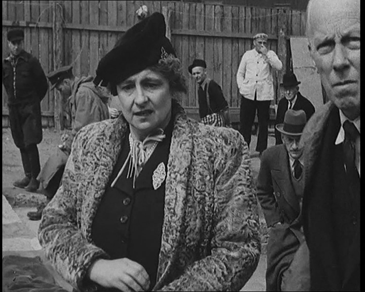 Eine historische Schwarz-Weiß-Fotografie aus dem Konzentrationslager Buchenwald 1945. Im Vordergrund steht eine Frau im Kostüm mit Pelzmantel und Hut, die ernst in die Kamera blickt. Rechts neben ihr ist ein älterer Mann teilweise zu sehen. Im Hintergrund befinden sich mehrere Männer, teils in Uniform, teils in Zivil, die auf den Boden schauen oder nachdenklich beiseite stehen. Die Szene vermittelt eine bedrückte, ernste Stimmung während des Besuchs einer Delegation, die die Verbrechen der Nationalsozialisten dokumentierte.