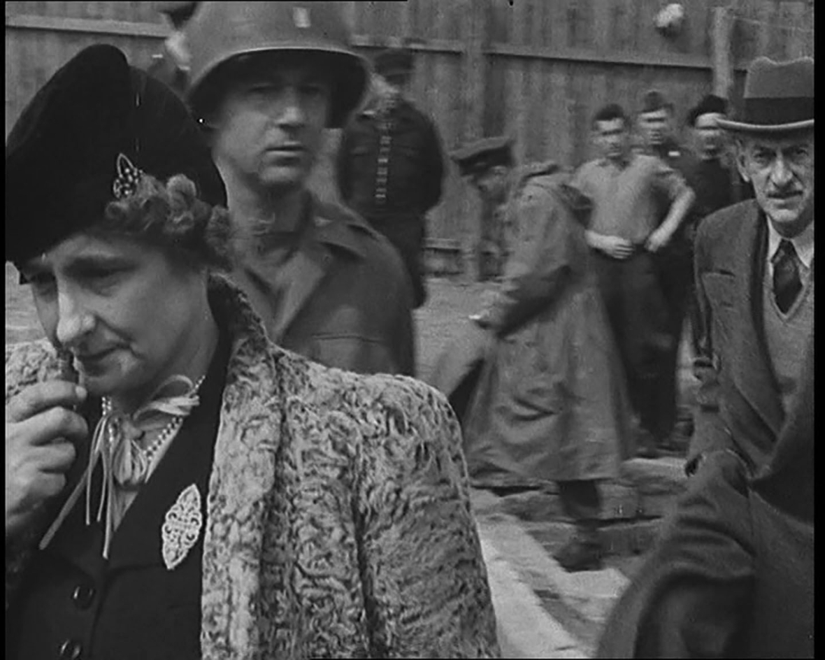 Eine historische Schwarz-Weiß-Fotografie aus dem Konzentrationslager Buchenwald 1945. Im Vordergrund steht eine Frau im Kostüm mit Pelzmantel und Hut, die ernst in die Kamera blickt. Rechts neben ihr ist ein älterer Mann teilweise zu sehen. Im Hintergrund befinden sich mehrere Männer, teils in Uniform, teils in Zivil, die auf den Boden schauen oder nachdenklich beiseite stehen. Die Szene vermittelt eine bedrückte, ernste Stimmung während des Besuchs einer Delegation, die die Verbrechen der Nationalsozialisten dokumentierte.