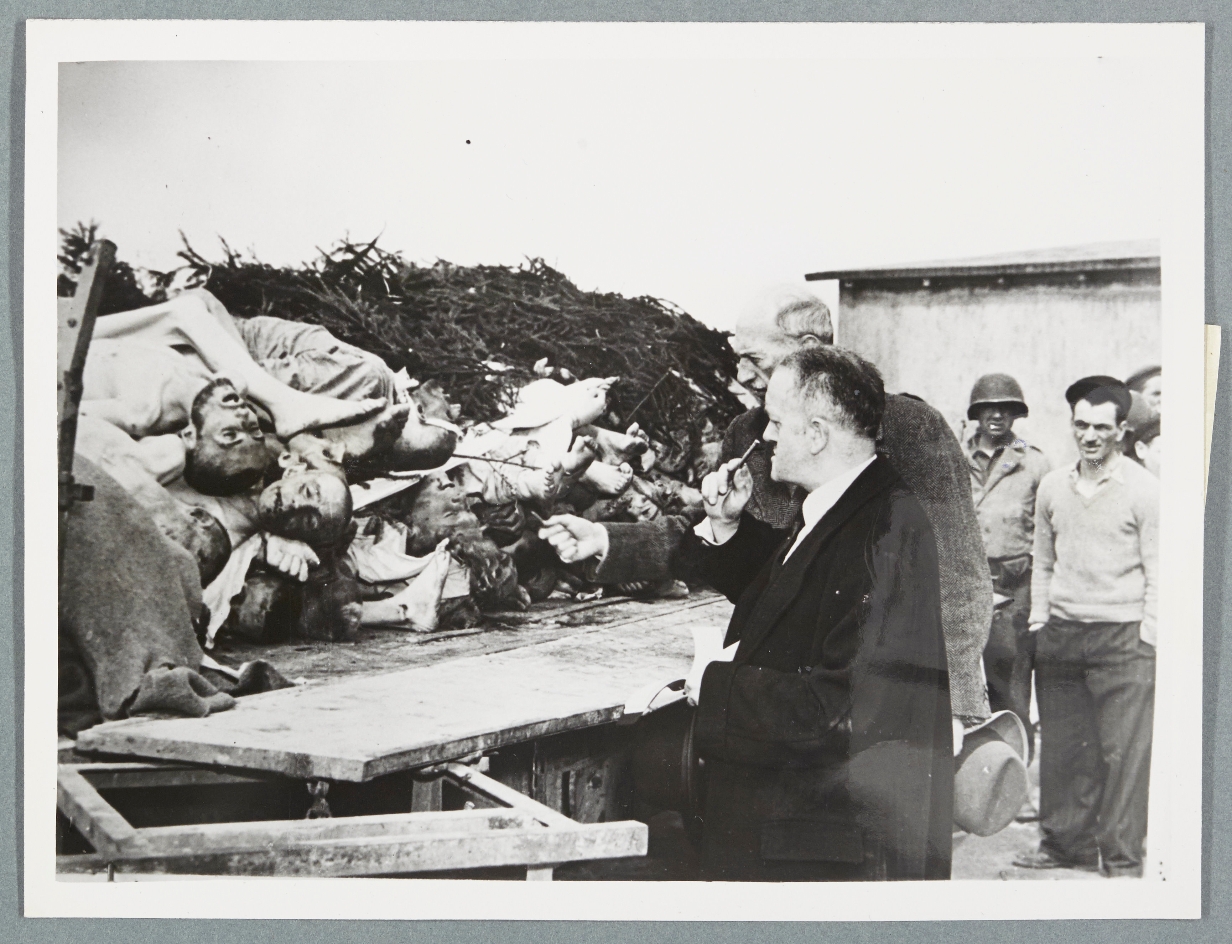 Eine historische Schwarz-Weiß-Fotografie zeigt Mitglieder einer Delegation beim Besuch des Konzentrationslagers Buchenwald 1945. Im Vordergrund begutachten zwei Männer in Zivilkleidung einen offenen Wagen, auf dem Leichen von KZ-Häftlingen übereinandergestapelt liegen. Einer der Männer hält nachdenklich eine Pfeife an den Mund. Im Hintergrund sind amerikanische Soldaten und weitere Personen zu sehen, die die Szene beobachten. Das Bild dokumentiert die unmittelbare Konfrontation mit den Verbrechen des NS-Regimes.