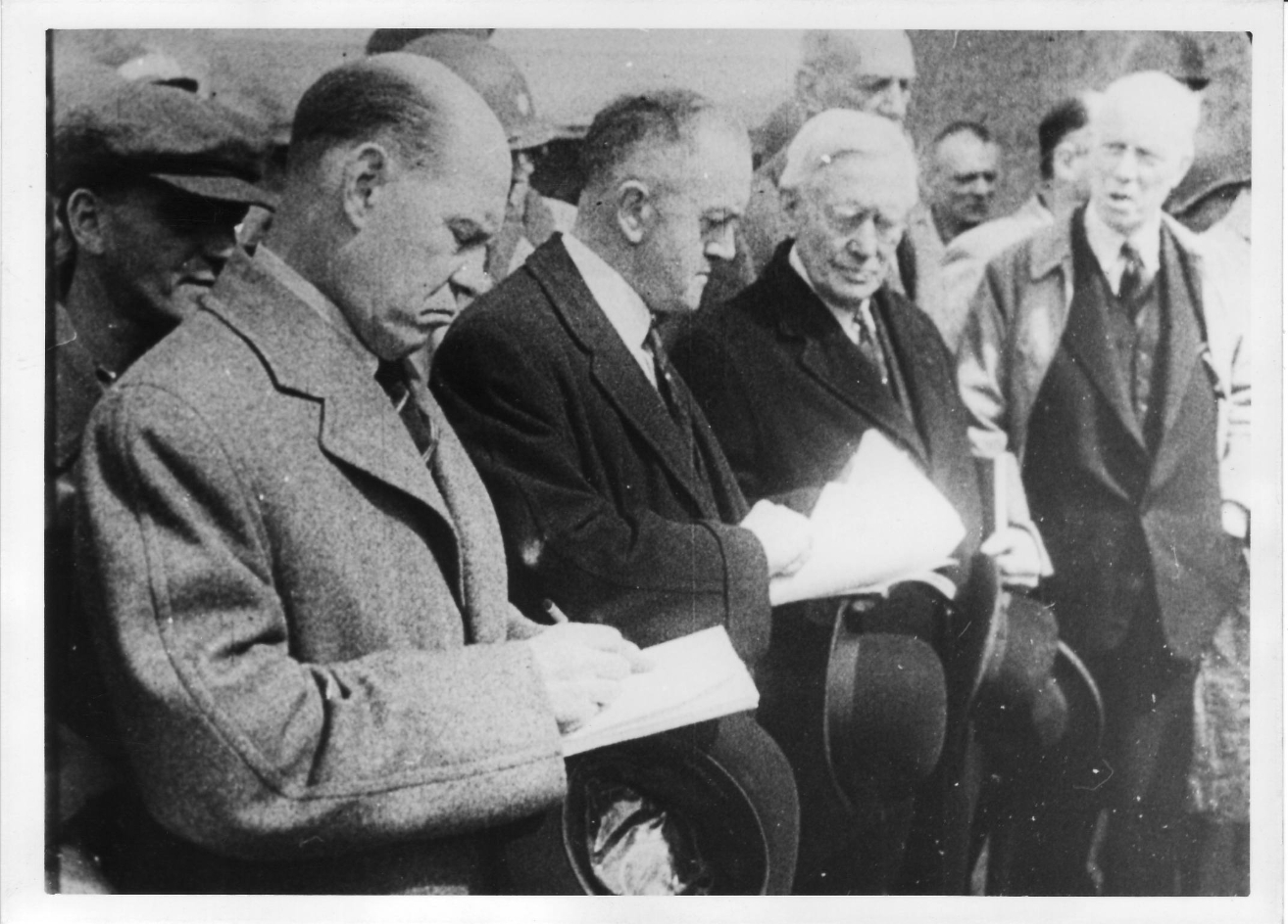 Eine historische Schwarz-Weiß-Fotografie zeigt mehrere Mitglieder einer britischen Parlamentsdelegation während ihres Besuchs im Konzentrationslager Buchenwald im April 1945. Im Vordergrund stehen drei Männer in Mänteln, die Notizen in Hefte schreiben, während sie ihre Hüte in der Hand halten. Neben und hinter ihnen sind weitere Männer zu sehen, darunter amerikanische Soldaten und andere Beobachter. Die Szene vermittelt eine ernste und konzentrierte Atmosphäre des Dokumentierens und Zeugenschaft-Ablegens.
