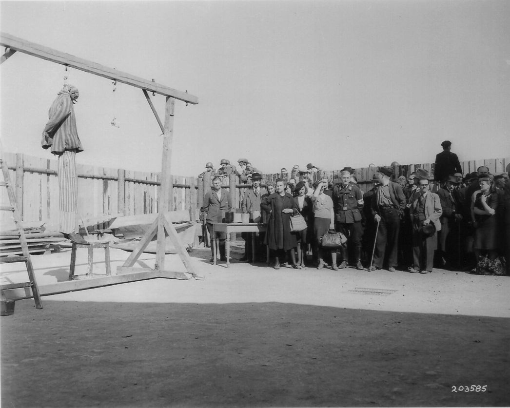 Auf dem Bild ist eine Gruppe weimarer Bürger zu sehen die neben einem Galgen im Krematoriumshof des KZ Buchenwalds stehen. Sie blicken auf den Galgen an dem zwei Stricke befesstigt sind. An dem Strick am linken Bildrand hängt eine Puppe in Häftlingskleidung.
