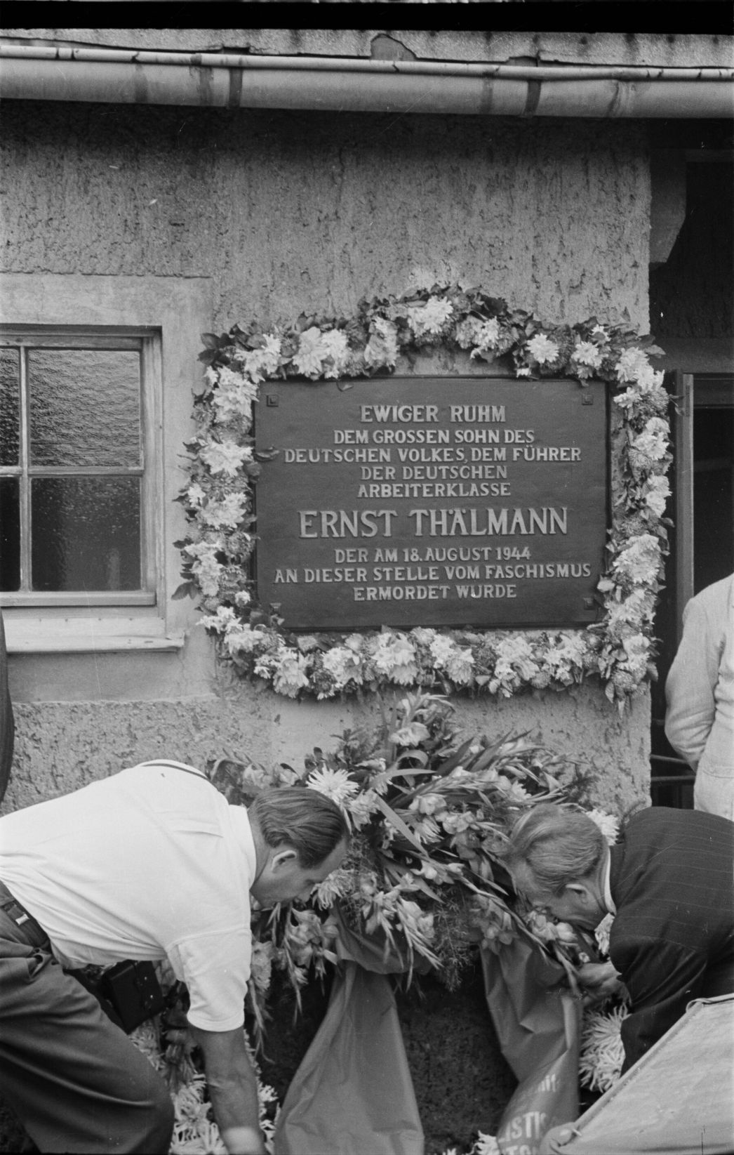 Zu sehen ist eine mit Blumen umrandete Gedenkstafel, die neben dem Eingang zu Krematorium hängt. Vor der Gedenktafel legen gerade zwei Männer einen großen Gedenkkranz ab.