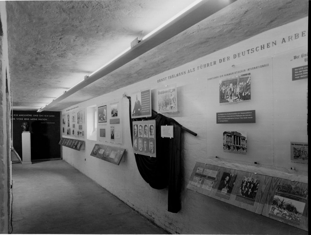 Zu sehen ist ein länglicher Gang an dessen rechter Wand verschidene Ausstellungstafeln und -Gegenstände angebracht sind. Am Ende das Gangs steht eine Büste, die den Kopf von Ernst Thälmann abbildet. 