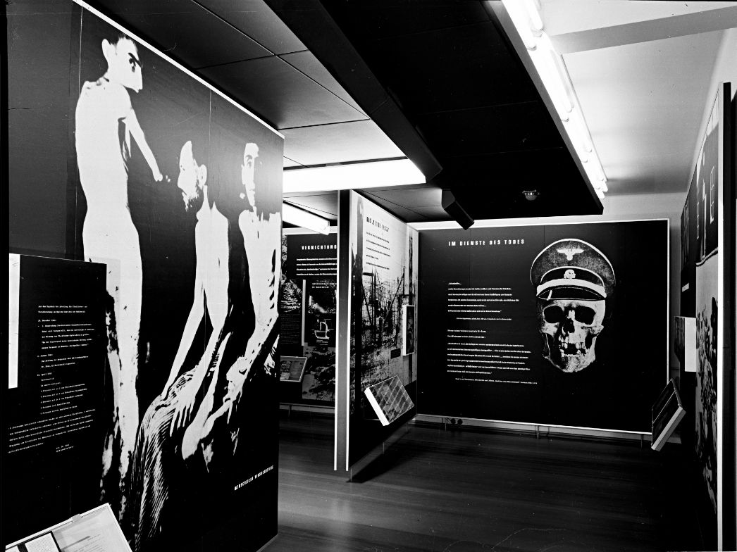 Zu sehen sind Ausstellungswände mit großen Abbildungen. Auf der linken Seite ist eine stark vergrößerte Fotografie von abgemagerten Häftlingen zu sehen. Auf der rechten Seite des Bildes ist ein Totenschädel mit SS-Schirmmütze zu sehen.