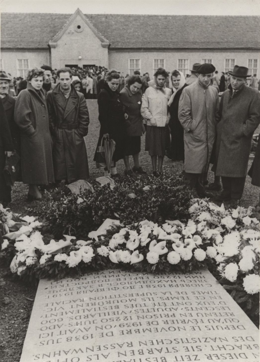 Zu sehen ist eine Gruppe von Menschen die sich vor einem Gedenkstein versammelt haben. Zu ihren Füßen sliegen um den Gedenkstein mehrere Blumenkränze und -Gestecke. Im Hintergrung ist die ehemalige Häftlingskantine zu sehen.