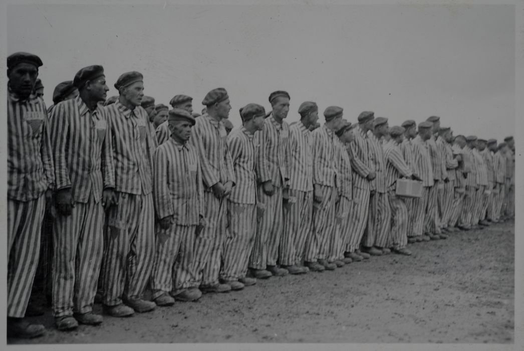 People dressed in striped, uniform prisoner clothing stand next to each other in narrow rows. Most of them look to the right. 
