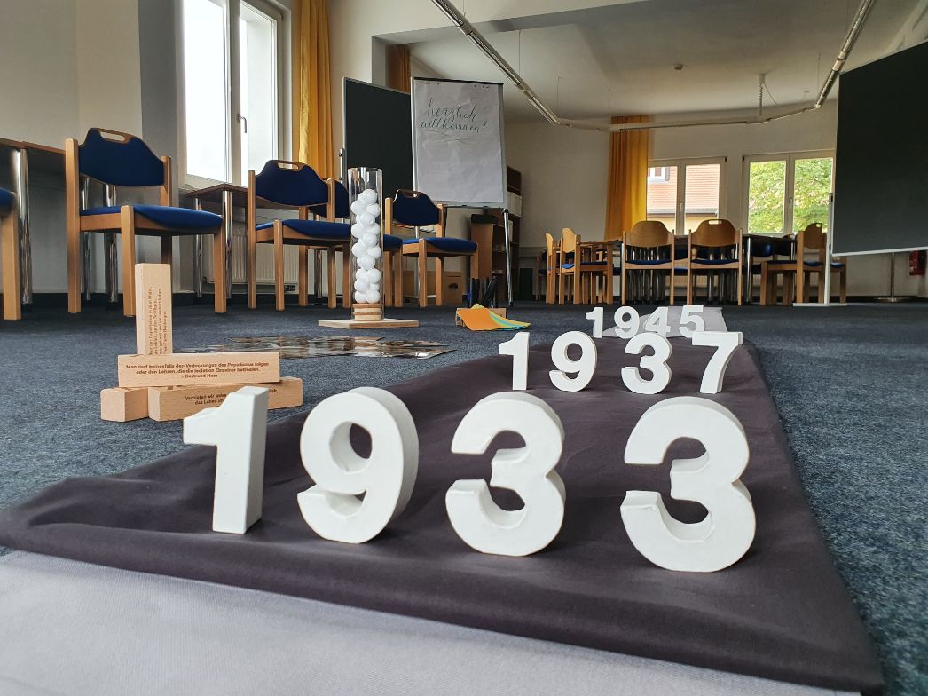 Blick in einen Seminarraum. Auf dem Boden liegt ein graues Tuch, darauf die Jahreszahlen 1933, dahinter 1937 und 1945. Im Hintergrund sind Stühle und Tische zu erkennen.
