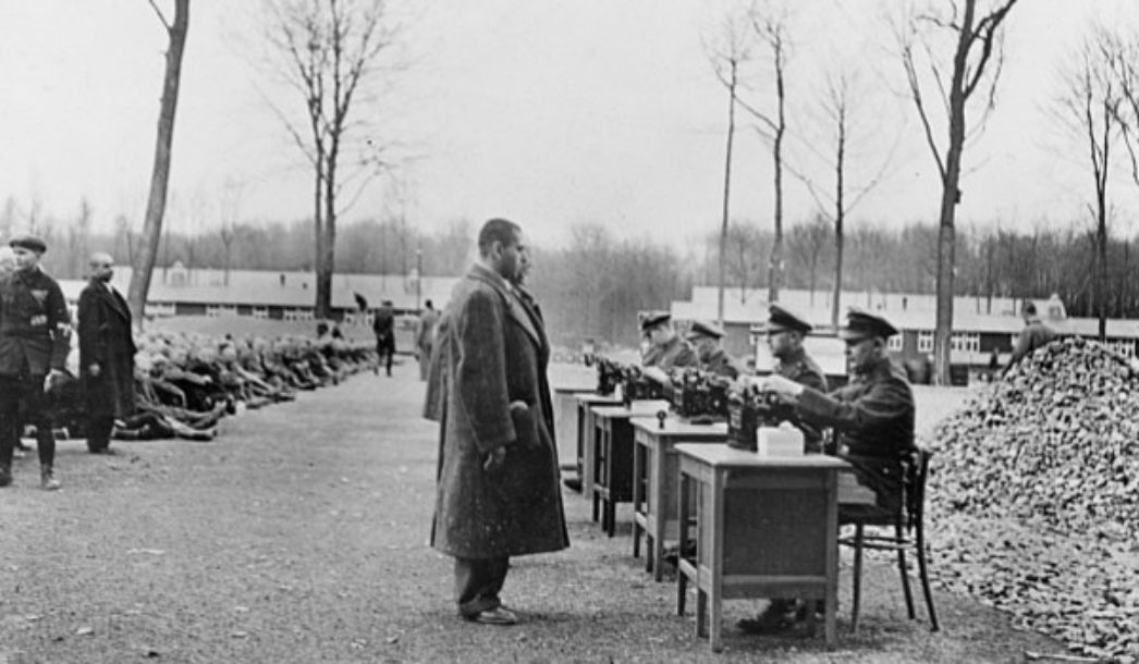 On the right side of the road, there are four tables outside at which uniformed people are sitting. People in civilian clothes are standing in front of the tables. On the left side of the path, numerous other people are sitting on the ground. In the background, flat functional buildings.
