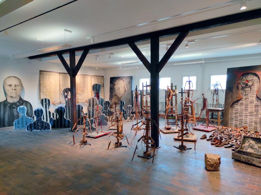 View of the installation Remineszenzen by Josef Szajna in the art exhibition of the Buchenwald Memorial. Arid, abstract wooden frames fill the space, vaguely reminiscent of people. In the background, larger-than-life portraits, a large list, and shoes on the floor. 