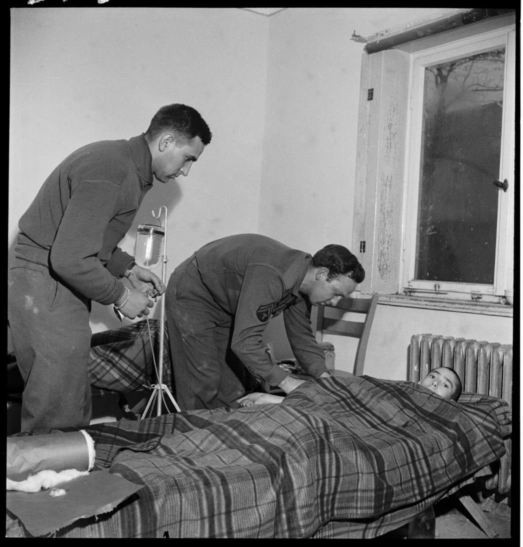 Auf dem Bild sind zwei Sanitäter der der US-Armee zu erkennen die links neben einer Pritsche mit einer befreiten Patientin stehen, die sich zur Patientin vorbeugen.