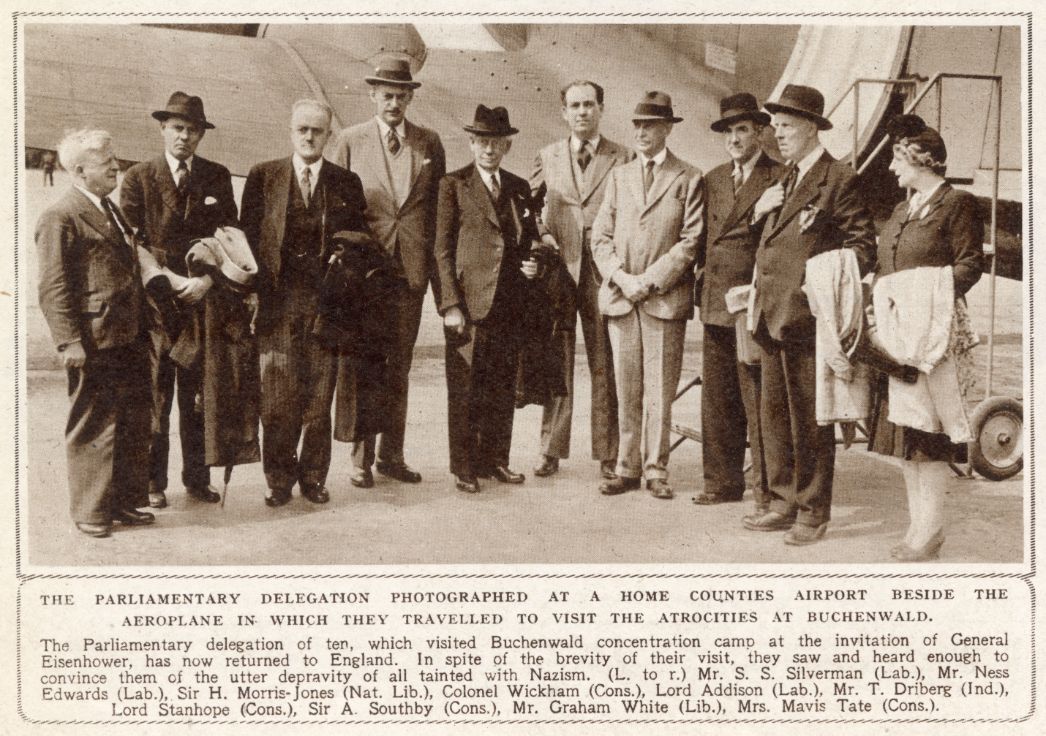 A black-and-white photograph of a group of ten people, mostly men in suits and hats, standing in front of an airplane. They are identified as a British parliamentary delegation that visited the Buchenwald concentration camp at the invitation of General Eisenhower. The group poses together, some holding coats and hats, with the airplane steps visible behind them. The caption lists their names and political affiliations, including S. S. Silverman, Ness Edwards, Sir H. Morris-Jones, Colonel Wickham, Lord Addison, T. Driberg, Lord Stanhope, Sir A. Southby, Graham White, and Mrs. Mavis Tate.