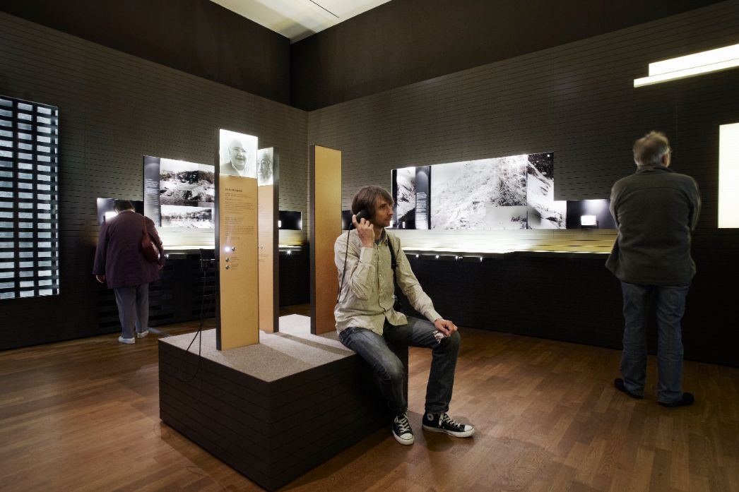 The picture shows the Forced Labour exhibition at the Jewish Museum in Berlin. In the center of the picture is a young man sitting on an island in the middle of the room. He is holding headphones to his ear. In the background, people can be seen facing the exhibition walls.