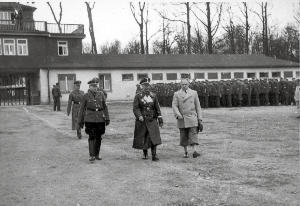 Schwarz-Weiß-Fotografie: drei uniformierte Männer vom Steinbruchkommando vorm Lagertor, im Hintergrund treten Häftlinge an