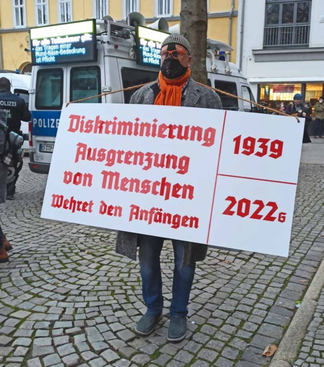 maskierter Mann hält großes Schild hoch, das den Nationalsozialismus mit Corona-Maßnahmen vergleicht