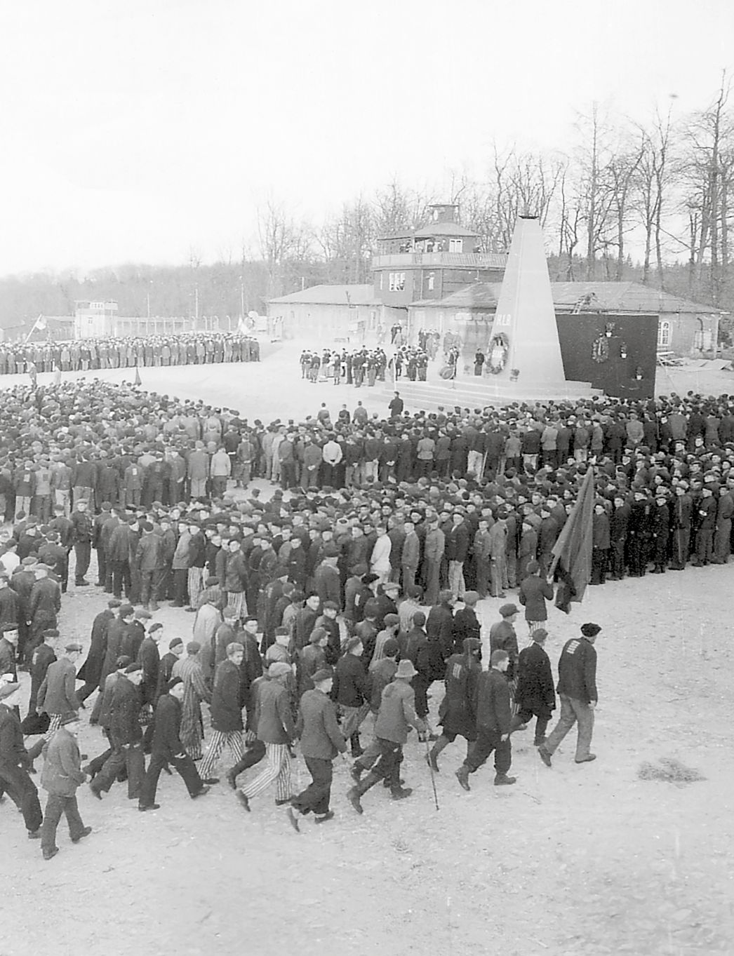 Schwarz-Weiß-Foto: Menschenmenge (ehemalige Häftlinge) vor dem Obelisken, der wiederum auf dem Lagergelände vor dem Lagertor steht